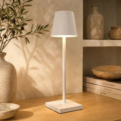 Lámpara LED portátil blanca encendida sobre una mesa de madera, junto a jarrones de cerámica y ramas de olivo